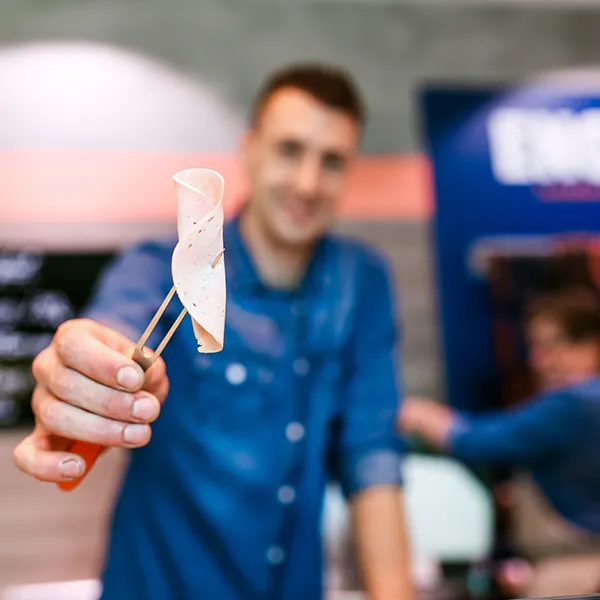 Mitarbeiter hält eine Scheibe Lyoner auf einem Spieß und reicht es über die Theke.